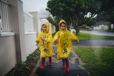 Fröhliche junge Mädchen in Regenmantel und Gummistiefeln laufen im Freien. Zwillingsschwestern in wasserdichten Mänteln an einem regnerischen Tag. - JLPSF19077
