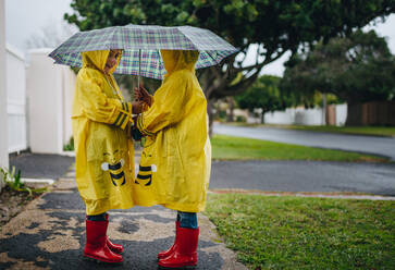 Zwei Kinder in wasserdichten Mänteln und roten Gummistiefeln stehen unter dem Regenschirm. Kleine Mädchen im Freien an einem regnerischen Tag. - JLPSF19078