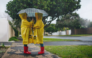 Zwei kleine Mädchen in wasserdichten Mänteln und Gummistiefeln stehen unter dem Regenschirm im Freien. Kleine Schwestern im Freien an einem regnerischen Tag. - JLPSF19080