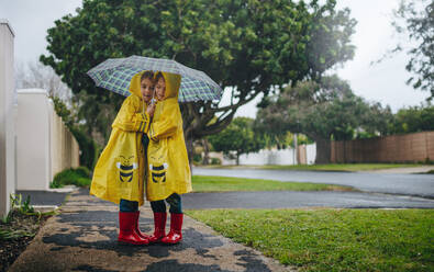 Two adorable little girls standing outdoors on a rainy day under an umbrella wearing raincoats. Twin sisters in waterproof coats standing together under umbrella outside. - JLPSF19081