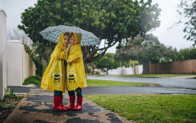 Zwei bezaubernde kleine Mädchen stehen an einem regnerischen Tag unter einem Regenschirm im Freien und tragen Regenmäntel. Zwillingsschwestern in wasserdichten Mänteln stehen zusammen unter einem Regenschirm im Freien. - JLPSF19081