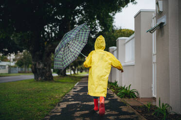 Rückansicht eines kleinen Mädchens, das an einem regnerischen Tag im Freien läuft. Ein Mädchen in Regenjacke und Gummistiefeln, das einen Regenschirm hält, läuft im Freien. - JLPSF19084