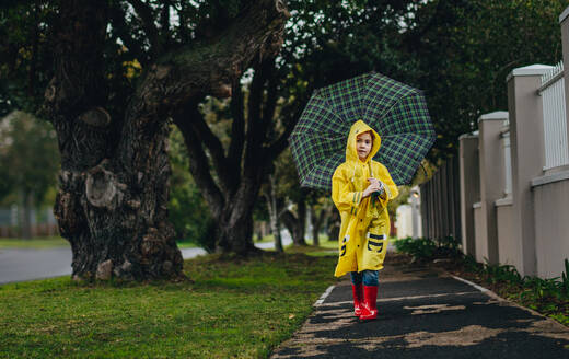 Porträt eines schönen jungen Mädchens, das an einem regnerischen Tag im Freien spazieren geht und dabei wasserdichte Kleidung und Gummistiefel trägt. Das Mädchen trägt einen Regenmantel und geht mit einem Regenschirm. - JLPSF19085