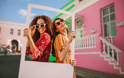 Two women wearing colourful dress and sunglasses standing outdoors holding a photo frame with houses in background. Beautiful female friends enjoying outdoors with a blank picture frame. - JLPSF19097
