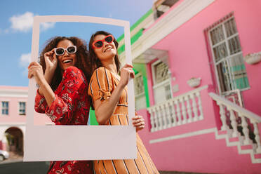 Two smiling woman standing together holding empty frame outdoors. Beautiful female travelers wearing sunglasses holding a blank picture frame. - JLPSF19098