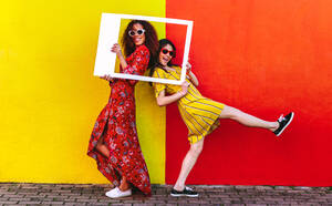 Two women friends with blank photo frame standing against colored wall outdoors. Female travelers posing at camera with empty picture frame. - JLPSF19100