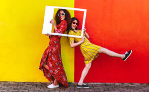 Zwei Freundinnen mit leerem Fotorahmen, die im Freien an einer farbigen Wand stehen. Weibliche Reisende posieren mit leerem Fotorahmen vor der Kamera. - JLPSF19100