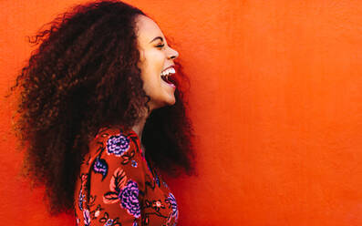 Seitenansicht einer fröhlichen Frau mit lockigem Haar vor orangefarbenem Hintergrund. Nahaufnahme einer lachenden afrikanischen jungen Frau. - JLPSF19112