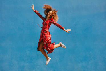 Young female model in red sundress jumping with joy. Beautiful young woman jumping over blue background. - JLPSF19115