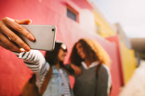 Woman taking selfie with female friend using mobile phone. Focus on smartphone in hand of woman used to making a self portrait outdoors with her friend. - JLPSF19118