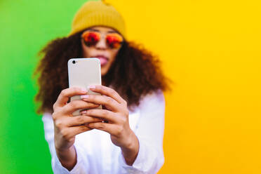 Stylish african girl taking a selfie using mobile phone. Blurred image of woman standing against coloured background and posing for a selfie. - JLPSF19129