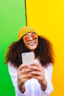 Schöne afrikanische Frau mit lockigem Haar, Hut und Sonnenbrille, die ein Selfie mit ihrem Handy macht. Mädchen steht vor einem farbigen Hintergrund und macht ein Selfie. - JLPSF19130