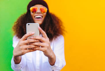 Curly haired african girl taking a selfie using smartphone. Focus on mobile phone in hand of woman taking selfie on yellow and green background. - JLPSF19131