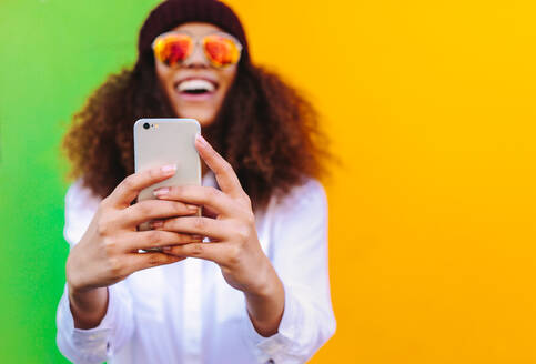 Lockenhaariges afrikanisches Mädchen, das ein Selfie mit einem Smartphone macht. Fokus auf das Handy in der Hand der Frau, die ein Selfie auf gelbem und grünem Hintergrund macht. - JLPSF19131