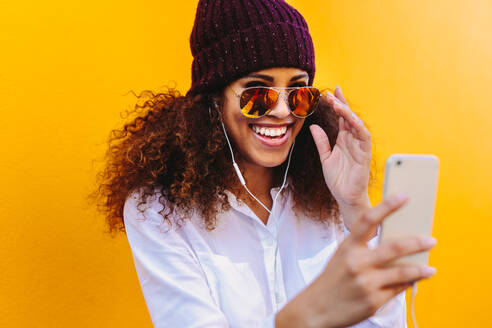 Schöne Frau, die ein Video-Chat mit Handy. Stilvolle Frau mit lockigem Haar trägt Hut und Sonnenbrille machen einen Videoanruf gegen gelben Hintergrund. - JLPSF19133