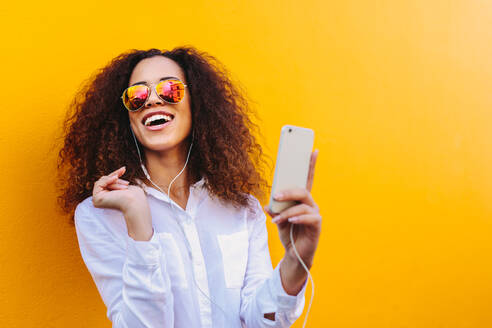 Glückliche junge afrikanische Frau, die mit einem Mobiltelefon Musik über Kopfhörer hört. Mädchen mit lockigem Haar, das eine Sonnenbrille trägt, vor einer gelben Wand, die Lieder auf einem Mobiltelefon hört. - JLPSF19134