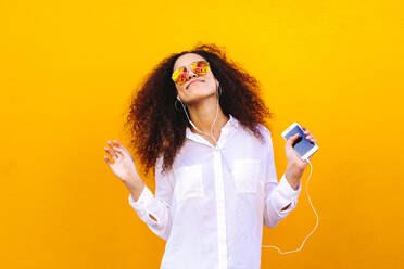African woman listening to music using earphones. Girl with curly hair wearing sunglasses and earphones listening to music from her smart phone on yellow wall. - JLPSF19135