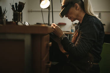 Seitenansicht einer älteren Frau mit Lupenbrille, die an ihrer Werkbank sitzt und das Produkt während des Herstellungsprozesses untersucht. - JLPSF19142