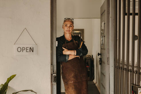 Portrait of senior female goldsmith standing at workshop door. Woman jeweler wearing apron and holding tools at workshop. - JLPSF19150