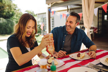 Lächelnder Mann und lächelnde Frau essen Hot Dogs am Esstisch. Frau gießt Tomatensoße in ihren Teller beim Essen. - JLPSF19169