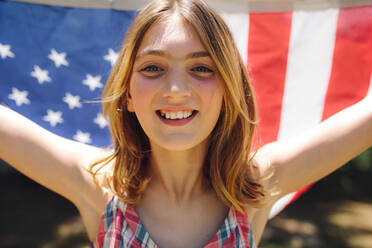 Nahaufnahme eines lächelnden Mädchens, das eine amerikanische Flagge hinter sich hält. Glückliches Mädchen, das im Freien steht und die amerikanische Flagge hält. - JLPSF19180