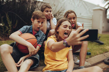 Kinder machen lustige Gesichter und strecken ihre Zunge für ein Selfie heraus. Ein Mädchen macht ein Selfie mit ihren Freunden im Freien. - JLPSF19193