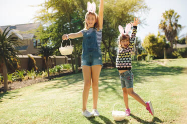 Zwei Kinder, die in einem Garten spielen und Körbe halten. Mädchen tragen ein schickes Kaninchenohr-Stirnband und spielen an einem sonnigen Tag im Freien. - JLPSF19198