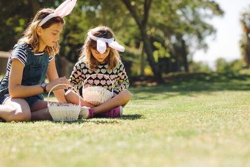 Zwei Mädchen, die ein Kaninchenohr-Stirnband tragen, spielen im Garten mit Körben. Kinder sitzen im Gras und spielen zusammen an einem sonnigen Tag. - JLPSF19200