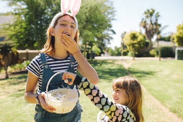 Kinder essen Süßigkeiten aus einem Korb, der in einem Garten steht. Kinder, die an einem sonnigen Tag in einem Garten spielen. - JLPSF19201