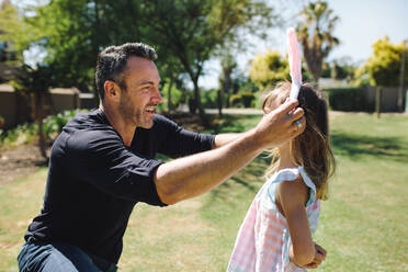 Vater setzt seiner kleinen Tochter in einem Park ein schickes Stirnband auf den Kopf. Vater und Tochter spielen an einem sonnigen Tag in einem Park. - JLPSF19205