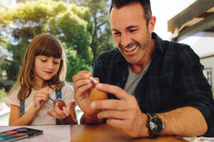 Lächelnder Mann, der Ostereier bemalt. Kleines Mädchen, das mit einem Pinsel Ostereier bemalt, sitzt mit ihrem Vater zusammen. - JLPSF19211