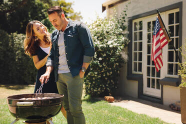 Lächelnder Mann und Frau kochen in ihrem Garten mit amerikanischer Flagge im Hintergrund. Paar macht gegrilltes Essen und feiert einen Anlass. - JLPSF19217