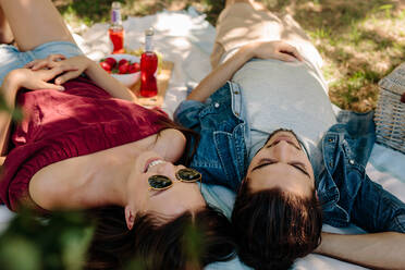 Junges Paar liegt auf einer Picknickdecke und lächelt. Frau mit Sonnenbrille entspannt sich mit ihrem Freund und lächelt. - JLPSF19224