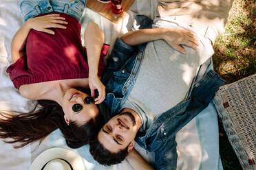 Shot from above of young couple in love lying on picnic blanket. Caucasian man with beard and woman wearing sunglasses relaxing together at the park. - JLPSF19225