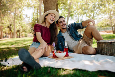 Fröhliches junges Paar, das im Park sitzt und ein Picknick macht. Lächelndes Paar, das sich zusammen mit einer Obstschale und Bier auf einer Picknickdecke entspannt. - JLPSF19226