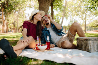 Young man offers a bit of strawberry to his girlfriend. Couple enjoying their time together and are having a picnic at park. - JLPSF19227