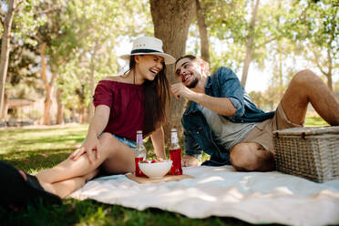 Lächelnder junger Mann und Frau sitzen auf einer Picknickdecke und haben Spaß. Fröhliches junges Paar genießt ein Picknick. - JLPSF19230