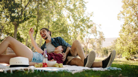 Ein lächelndes Paar macht ein Selfie beim Picknick. Eine Frau liegt auf dem Schoß ihres Freundes und macht ein Selfie mit ihrem Mobiltelefon. - JLPSF19237