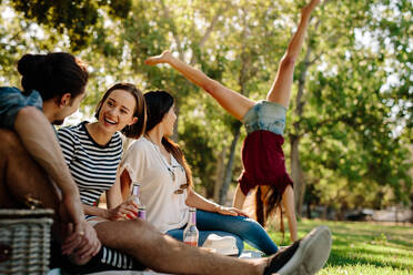 Eine Gruppe von Freunden sitzt im Park mit einer Frau, die im Hintergrund ein Rad schlägt. Junger Mann und Frauen genießen einen Sommertag im Park. Freunde genießen ein Picknick. - JLPSF19251