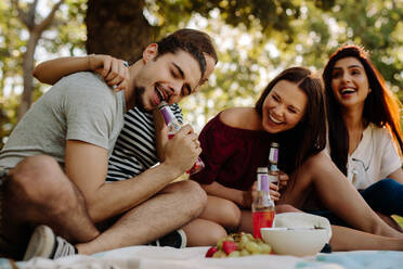 Ein Mann öffnet eine Flasche mit seinen Zähnen und eine Frau sitzt daneben und lächelt. Eine Gruppe von Freunden genießt ihre Getränke bei einem Picknick. - JLPSF19253