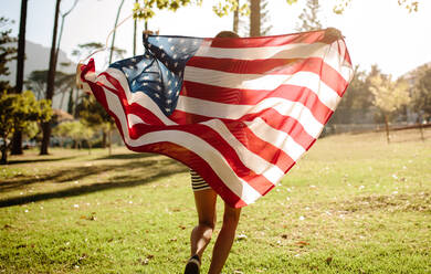 Rear view of young woman running in the forest with American Flag. American girl celebrating fourth of july in the park. - JLPSF19259