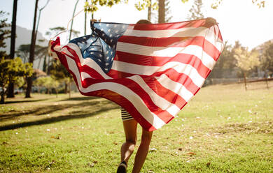 Rückansicht einer jungen Frau, die mit einer amerikanischen Flagge durch den Wald läuft. Amerikanisches Mädchen feiert den vierten Juli im Park. - JLPSF19259