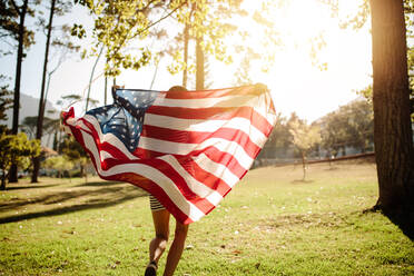 Frau läuft mit amerikanischer Flagge an einem sonnigen Tag. Schuss von hinten von einem Mädchen, das im Park mit amerikanischer Flagge läuft. - JLPSF19260