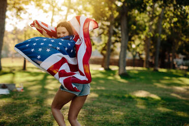 Junge patriotische Frau mit amerikanischer Flagge im Park schaut zurück und lächelt. Mädchen läuft mit USA-Flagge im Park. - JLPSF19261