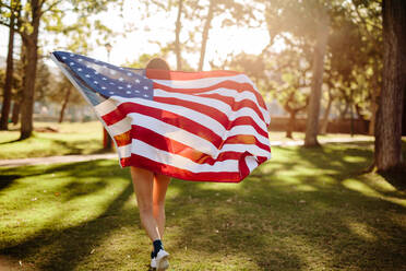 Junge Frau hält amerikanische Flagge und läuft im Park. Mädchen trägt amerikanische Flagge, während sie durch einen Park an einem Sommertag läuft. Feiern des vierten Juli. - JLPSF19262