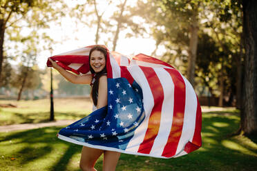 Patriotic girl with american flag in the park. Beautiful woman running with a USA flag outdoors at park. - JLPSF19263