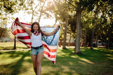 Beautiful young woman walking in the park and waving American flag. Smiling girl with American flag at park. - JLPSF19264