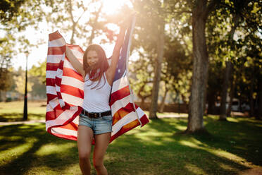 Attraktives Mädchen in Shorts läuft mit amerikanischer Flagge in den Händen. Lächelnde junge Frau mit USA-Flagge im Park. - JLPSF19265