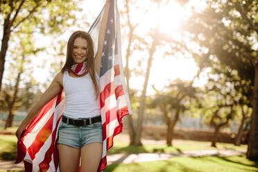 Portrait of a beautiful woman in the park with American flag. Smiling american girl holding national flag proudly and looking at camera. - JLPSF19266