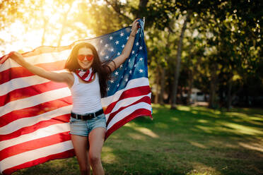 Mädchen in Shorts läuft im Park und hält amerikanische Flagge. Lächelndes Mädchen mit amerikanischer Flagge im Park am vierten Juli. - JLPSF19267